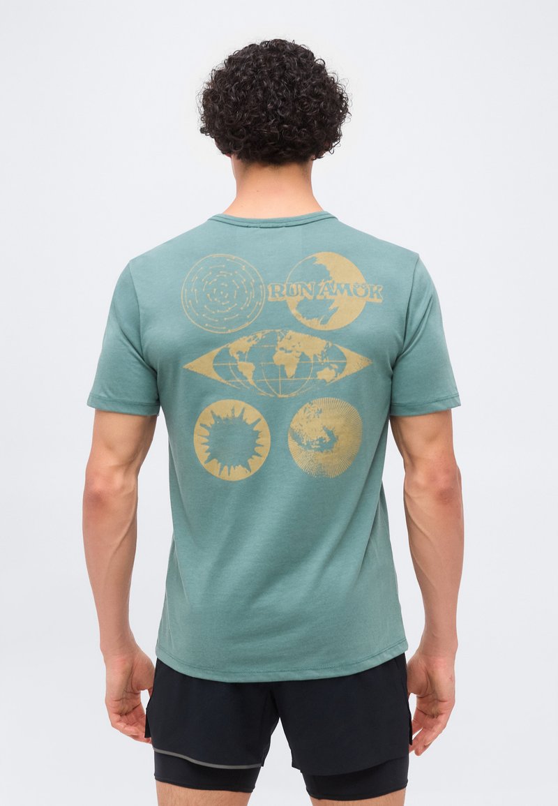 Άνδρας με σγουρά μαλλιά που φοράει τιρκουάζ μπλουζάκι με ξεθωριασμένο κίτρινο σχέδιο παγκόσμιου χάρτη, ήλιου, φεγγαριού και κυκλικών σχεδίων στην πλάτη, σε συνδυασμό με μαύρα σορτς.