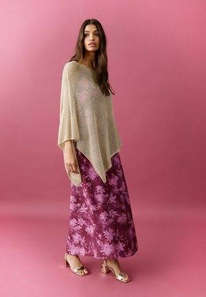 Ženska z dolgimi rjavimi lasmi, oblečena v bež pleteno pončo in vijolično žametno krilo s cvetličnim vzorcem na roza ozadju.