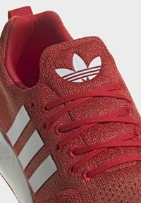 Röd idrottssko tillverkad av stickat material, med vit tre-stripes design, röda snören och en vit logotyp på tungan.