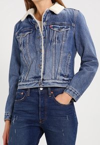 Giacca di jeans blu con collo foderato in shearling, tasche frontali e chiusure con bottoni. Texture consumata e design corto. Indossata con jeans scuri.