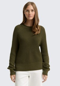 Oliwkowy sweter dzianinowy z prążkowaną fakturą, okrągłym dekoltem i luźnymi rękawami. Wykończony prążkowanym dołem, noszony na białych spodniach.