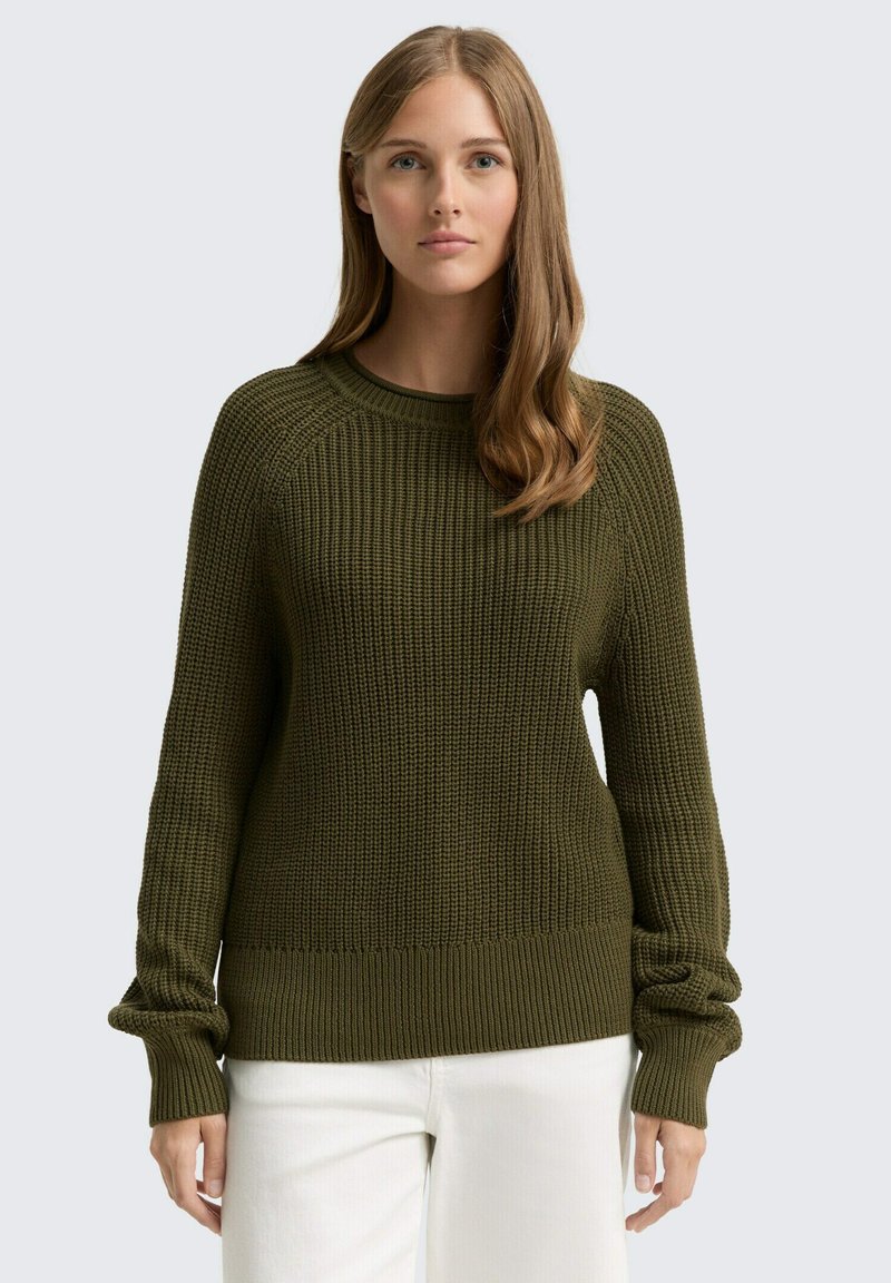 Oliwkowy sweter dzianinowy z prążkowaną fakturą, okrągłym dekoltem i luźnymi rękawami. Wykończony prążkowanym dołem, noszony na białych spodniach.