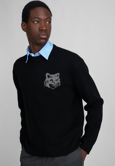 Mann in schwarzem Pullover mit grauem Wolfskopf-Emblem über blauem Hemd, graue Hose tragend, schaut zur Seite mit den Händen in den Taschen.