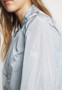 Ανοιχτό μπλε αδιάβροχο μπουφάν με λεία υφή, διαθέτει λάβαρο με logo στο αριστερό μανίκι και κλείσιμο με φερμουάρ μπροστά.