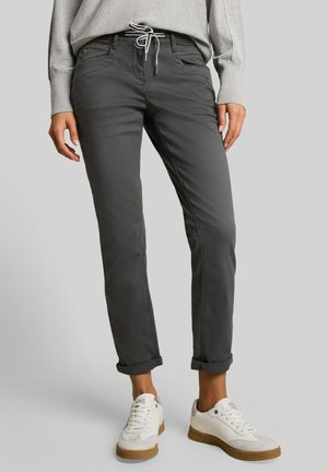 Personne portant un pantalon décontracté gris foncé avec des revers et des baskets blanches, debout sur un fond uni gris clair.