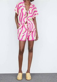 Ensemble chemise à manches courtes à boutons et short couleur crème avec des motifs audacieux en forme de vagues roses. Associé à des chaussures jaunes à enfiler.