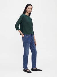 Femme aux longs cheveux noirs portant un haut texturé vert foncé, un jean bleu et des ballerines noires pointues, debout devant un fond blanc uni.