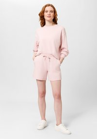Světle růžová mikina s dlouhými rukávy a volným střihem, kombinovaná s odpovídajícími šortkami s vysokým pasem. Obsahuje kapsy a stahovací pas.