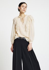 Cremefarbene Bluse mit Spitzenvolants, einem Schlüsselloch-Ausschnitt und Ballonärmeln, kombiniert mit hoch taillierten schwarzen plissierten Hosen und einem Gürtel.