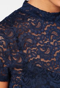 Navyblå spetsblus med intrikata blommönster, en texturerad yta och en volangkrage. Genomskinligt tyg med korta ärmar.