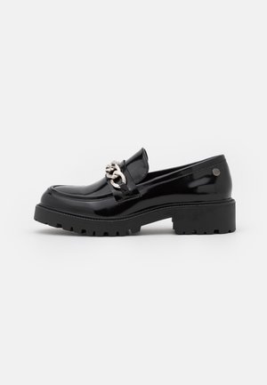 Mocassin noir brillant avec semelle crantée épaisse et détail de chaîne argentée sur le dessus, présenté sur fond blanc.