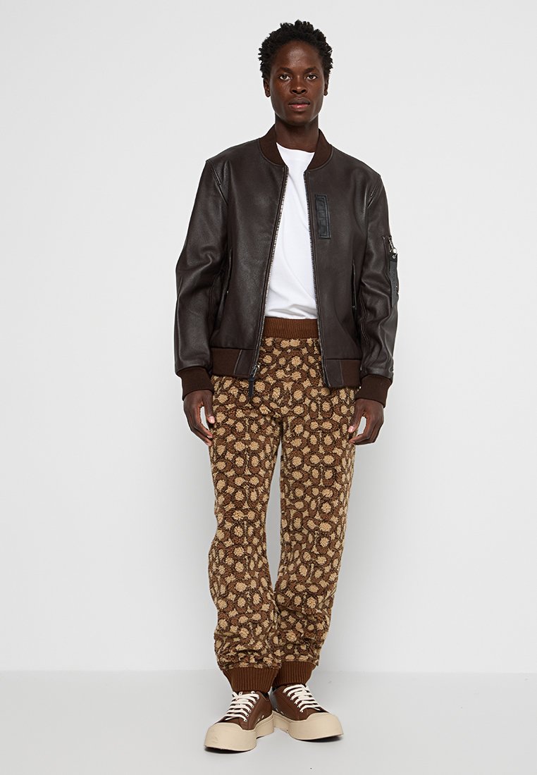 Brązowa skórzana kurtka bombowa z prążkowanymi mankietami, biała koszulka, wzorzyste brązowe spodnie z kwiatowym wzorem i kremowe sneakersy.