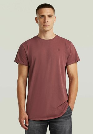 Uomo con maglietta bordeaux a maniche arrotolate con piccolo logo sul petto e jeans grigio scuro, in piedi contro uno sfondo chiaro e uniforme.