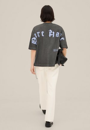 Henkilö, jolla on lyhyet tummat hiukset, kävelee poispäin. Hänellä on yllään tumma, ylikokoinen T-paita, jossa on iso vaaleansininen teksti, sekä valkoiset housut. Hän pitää kädessään mustaa laukkua.