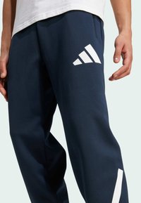 Granatowe spodnie sportowe wykonane z gładkiego materiału, z białymi paskami adidas na lewej udzie i zwężającym się krojem.