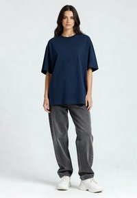 Tamsiai mėlynas didelių išmatavimų medvilninis marškinėlis su apvaliu kaklu, derinamas su pilkais džinsiniais kelinais ir baltais sportbačiais, stovintis neutralioje fone.