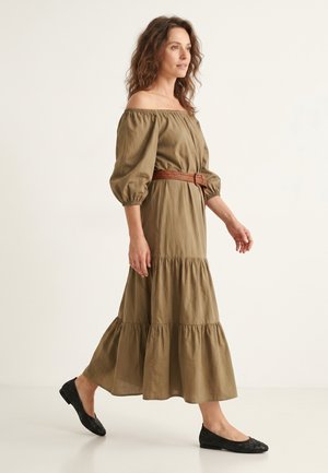 Vrouw die loopt in een olijfgroen off-shoulder jurk met pofmouwen, een bruine geweven riem en zwarte geweven platte schoenen tegen een effen achtergrond.