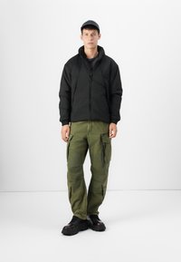 Doudoune noire avec un col monté, pantalon cargo vert avec plusieurs poches, et des bottes à lacets noires à semelle épaisse.