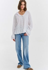 Kobieta ubrana w białą, długorękawową bluzkę z falbankami oraz jasnoniebieskie jeansy o prostych nogawkach, do tego wsuwane buty w panterkę.