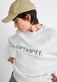 Grijze crewneck sweatshirt met blauwe branding, voorzien van ribbels aan de manchetten en de zoom. Beige pet met een textuur afgebeeld vanuit een hoek.
