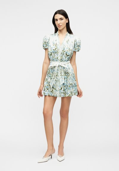 Woman wearing a short, floral-patterned dress with puffed sleeves and a tied belt, paired with white pointed-toe heels, standing against a plain background.