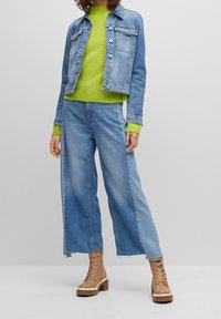 Chaqueta de denim con botones plateados, combinada con un suéter de canalé verde brillante y pantalones vaqueros de pierna ancha azules. Botines de cordones en marrón completan el conjunto.