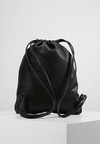 Sac à dos en cuir noir avec cordon de serrage et fines bretelles, posé sur une surface blanche devant un fond gris clair uni.
