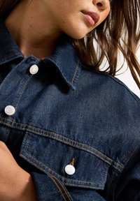 Mörkblå denimjacka med spetsig krage, två framfickor, silverknappar och uttalade sömdetaljer. Slät struktur.