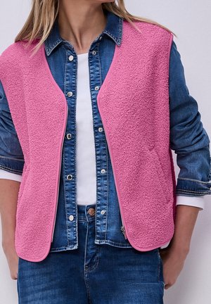 Vrouw draagt een roze gestructureerd mouwloos vest over een blauw denim overhemd met knopen en een wit topje, handen langs de zij.