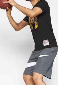 Maglietta nera da basket con un logo giallo e viola, abbinata a pantaloni sportivi grigi con dettagli bianchi e una superficie texturizzata.
