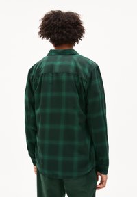 Persona con capelli ricci che indossa una camicia a maniche lunghe a quadri verde scuro e pantaloni verdi, in piedi di spalle su uno sfondo bianco.