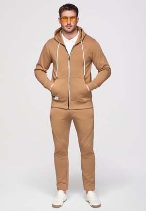 Mann steht mit den Händen in den Taschen, trägt einen hellbraunen Zip-Hoodie, passende Hose, weiße Turnschuhe, weißes Poloshirt und gelb getönte Brille.