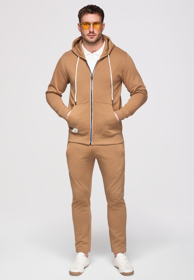 Homme debout les mains dans les poches, portant un sweat à capuche beige zippé, un pantalon assorti, des baskets blanches, un polo blanc et des lunettes à verres jaunes.