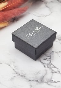Schwarze, strukturierte Schmuckbox mit Deckel, auf der weißen Schriftzüge auf der Oberseite zu sehen sind, platziert auf einer marmorierten Fläche.