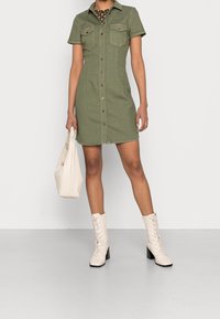 Donna che indossa un vestito a camicia verde oliva con maniche corte, una maglietta a righe marrone sotto, stivali bianchi alla caviglia e tiene in mano una borsa bianca.