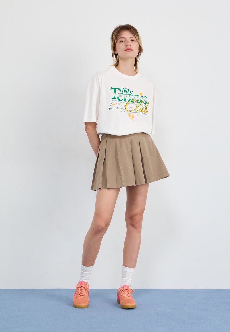 Weißes übergroßes T-Shirt mit grünem und orangefarbenem Logo, kombiniert mit einem braunen Faltenrock. Orangefarbene Turnschuhe ergänzen weiße Socken. Einfacher Hintergrund.