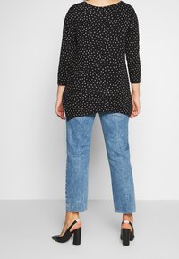 Túnica negra con lunares blancos, mangas de tres cuartos y un dobladillo más largo en la parte de atrás, combinada con unos vaqueros de mezclilla azul claro y zapatos de tacón bloque negro.