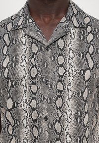 Chemise à manches courtes en imprimé serpent noir et beige, avec un col, une fermeture à boutons, et un tissu texturé avec des motifs variés.