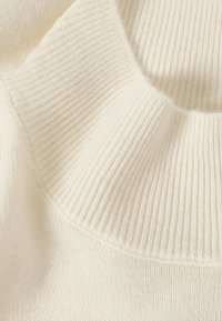 Tissu en maille de couleur crème avec un col montant côtelé. La texture est douce avec une finition lisse et des variations subtiles dans le tricot.