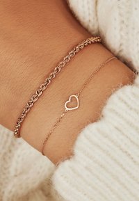 Twee roségouden armbanden om een pols; de ene met een gedraaide kettingontwerp, de andere een delicate ketting met een hartbedel. Zachte textuur en warme kleur.