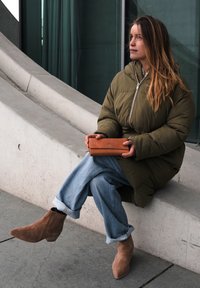 Olivgrüne Steppjacke, hellblaue weite Jeans und braune Wildleder-Stiefeletten. Braune Lederbrieftasche in den Händen gehalten. Betonhintergrund.