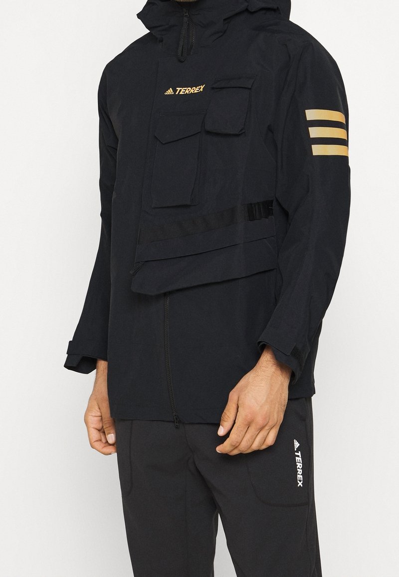 Veste à capuche noire avec une fermeture éclair sur le devant, plusieurs poches et des accents dorés. Le matériau a une texture lisse avec un design fonctionnel et superposé.