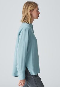 Lichtblauwe blouse met lange mouwen, een staande kraag, zijsplitten en knoopdetails op de manchetten. Zachte stof met een gladde textuur.