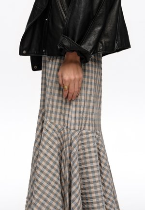 Mano con anillos de oro descansando al lado, vistiendo chaqueta de cuero negra y falda acampanada a cuadros beige y azul, sobre fondo blanco.