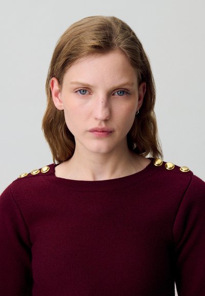 Maglione a maglia bordeaux con dettagli in bottone dorato sulle spalle, scollatura tonda e tessuto testurizzato.