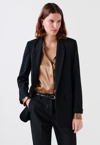 Musta räätälöity bleiseri, jossa on slim fit -leikkaus, yhdistettynä silkkiseen vaalean kultaisiin puseroon. Varustettu taskulla ja mustalla vyöllä, jossa on kultainen solki.