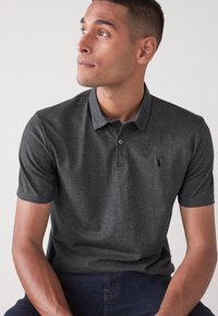 Polo de color gris oscuro con mangas cortas, cuello y un sutil detalle de logo. Hecho de una tela texturizada. Modelo sentado, vistiendo jeans azul marino.