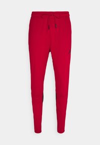 Pantalones deportivos rojos con cintura ajustable, piernas estrechas, acentos laterales negros y un pequeño logo negro en el muslo izquierdo. Textura de tela suave.