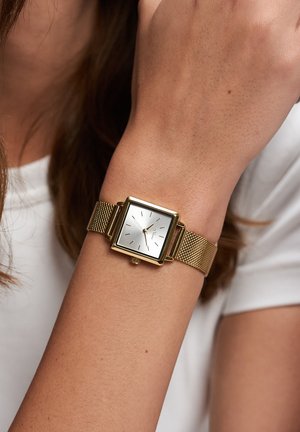 Guld firkantet ur med sølvfarvet urskive, minimalistiske timemarkeringer og meshrem, båret på et håndled med en lysefarvet skjorte i baggrunden.