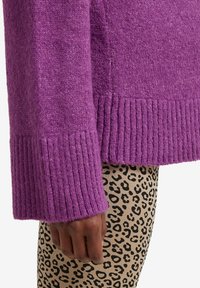 Dunkelhäutige Hand in der Nähe der Hüfte, die einen lila Strickpullover über beigen Leggings mit schwarzen Leopardenpunkten vor weißem Hintergrund trägt.